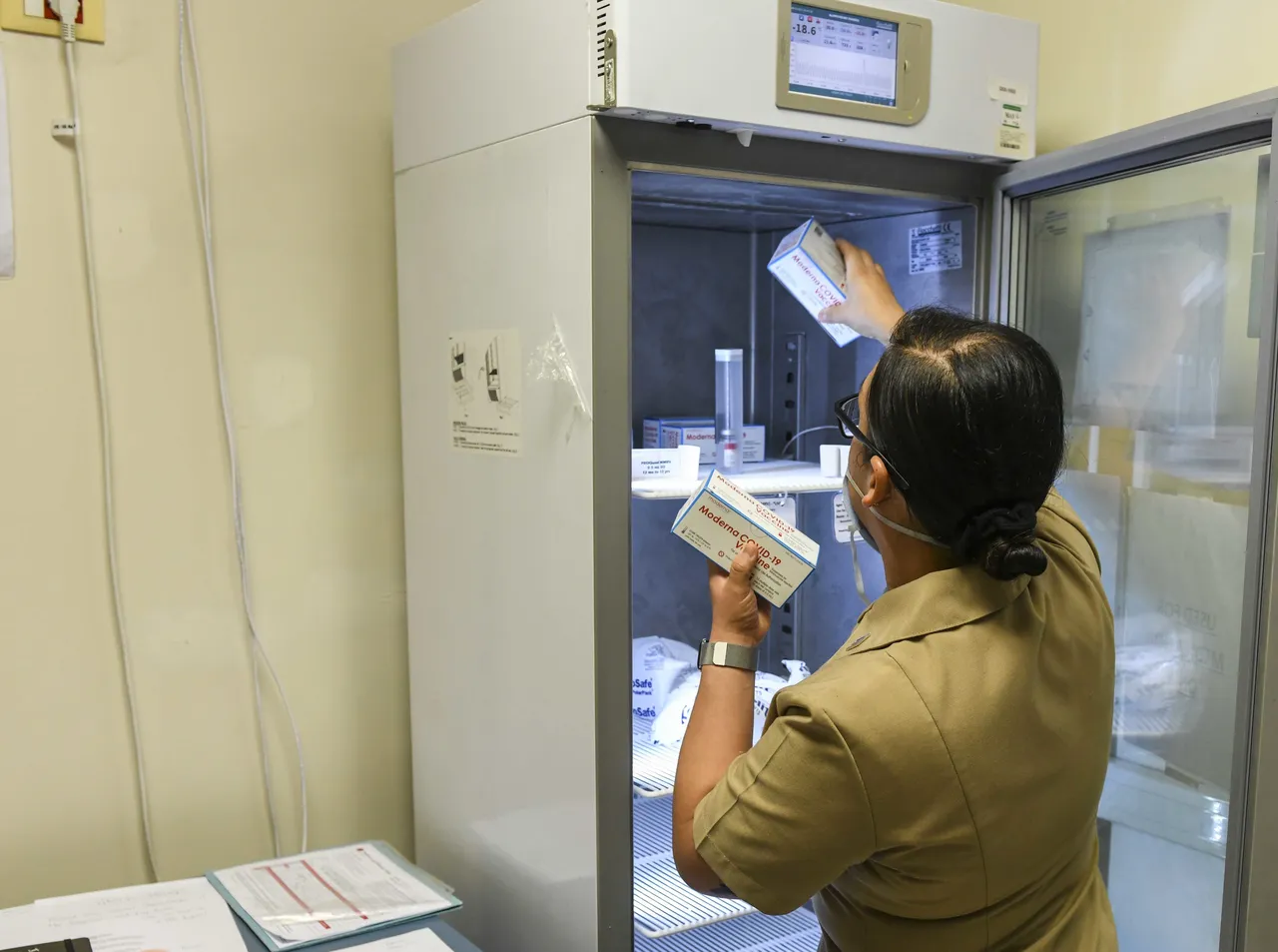 A hospital worker stocking vaccine boxes in a medical freezer, its temperature on the display.